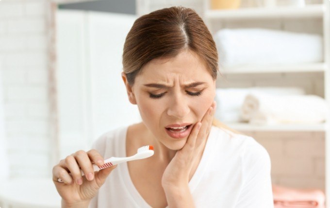 Woman holding toothbrush in one hand and touching her jaw in pain with the other hand